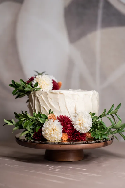 Cream-frosted wedding cake topped with burgundy and white dahlias and green foliage on a wooden cake stand.