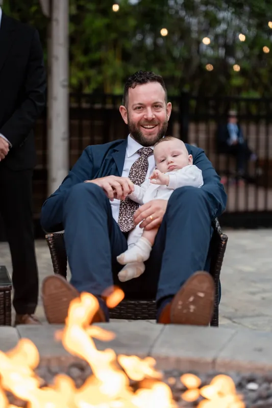 Man in navy suit holding an infant in white while seated by a firepit under string lights at Brick and Beam, Jacksonville.