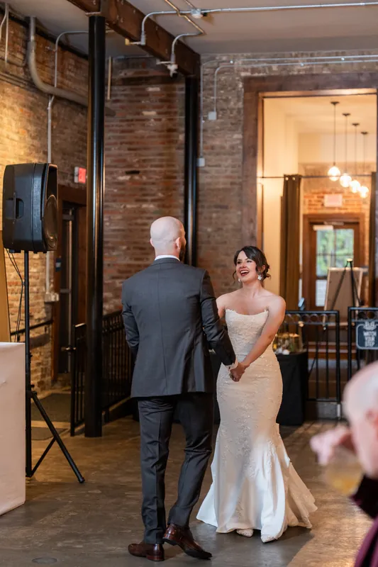 Two people in wedding attire face each other in a brick hallway with warm overhead lighting and guests nearby.
