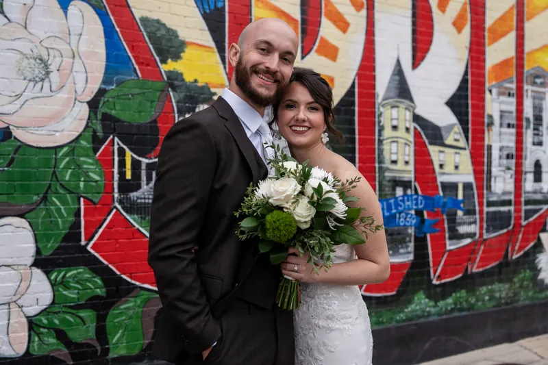 Faith and Christian pose together at Brick and Beam, holding a white and green bouquet in front of colorful mural art.