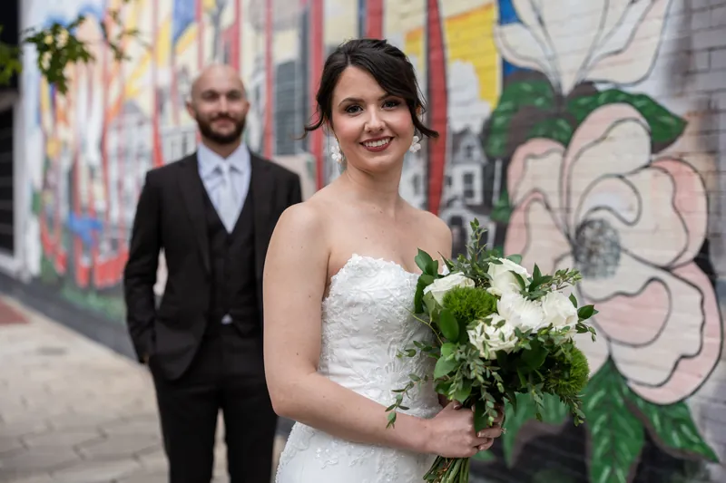 Someone in a white lace gown holds greenery while standing before street art, with a partner in a dark suit visible behind.