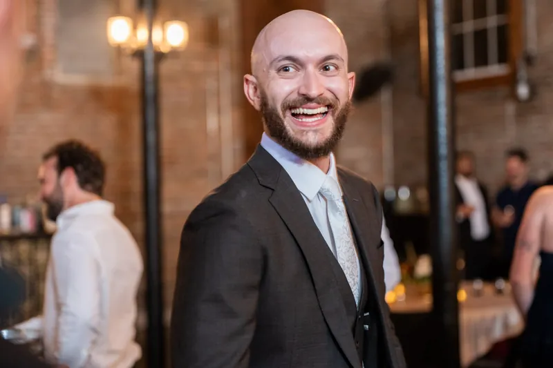 A bearded man in a charcoal suit and light blue shirt grins at an indoor reception, guests mingling in the background.