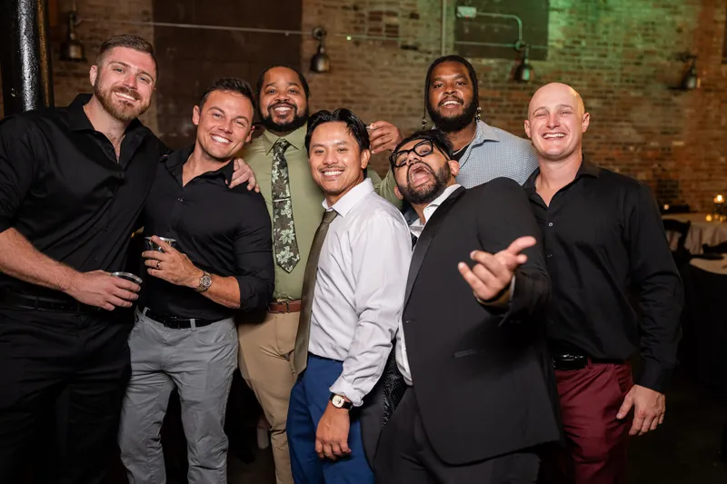 Six male guests pose shoulder-to-shoulder indoors, laughing at the reception.