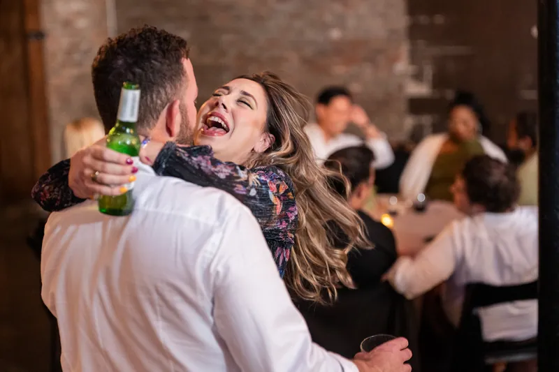 Two people dance together, one holding a beer bottle, surrounded by other guests at the reception.