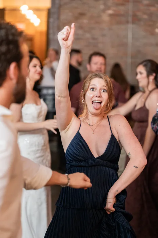 Woman in a navy dress raises her fist mid-celebration at Brick and Beam, surrounded by dancing guests in warm ambient light.