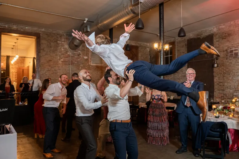 Guests toss someone in a light blue shirt skyward during an energetic dance moment at Brick and Beam's brick-lined reception hall.