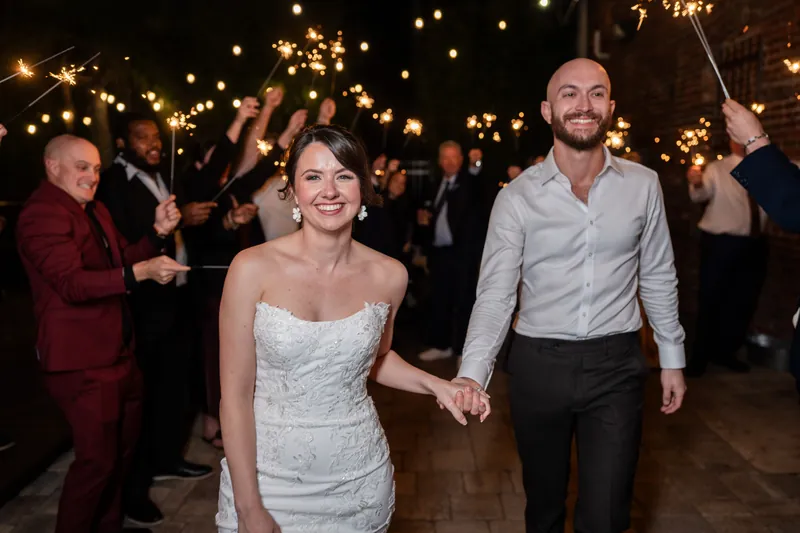 Faith and Christian walk hand-in-hand under a canopy of string lights as guests line the aisle at Brick and Beam's outdoor exit.