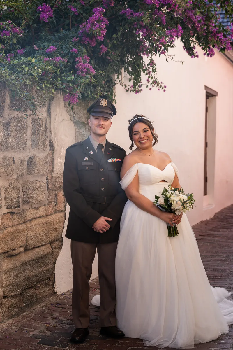 Gabi holds a white bouquet while Dawson wraps his arm around her waist against a pink-stuccoed building downtown.
