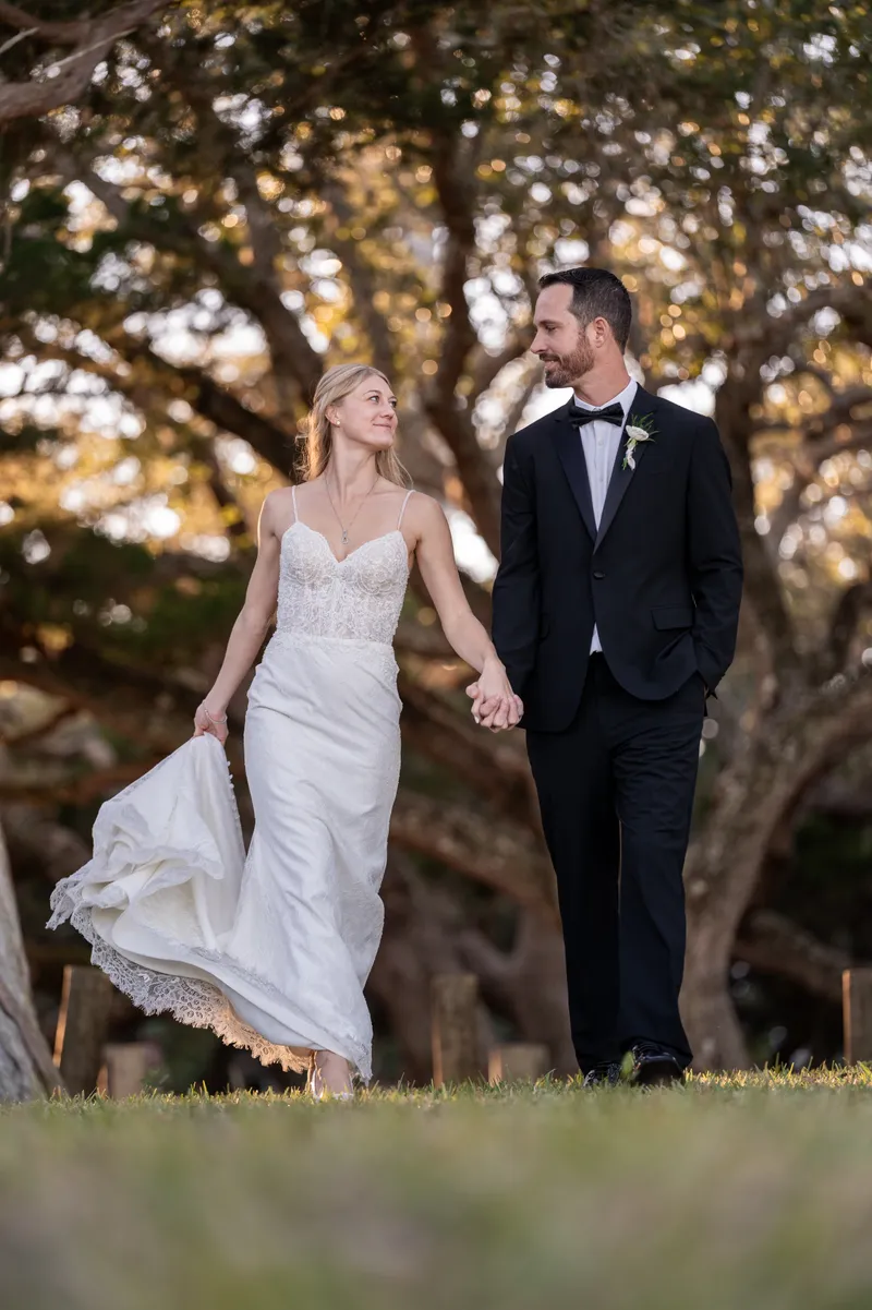 Bride and groom holding hands walking under large trees at golden hour