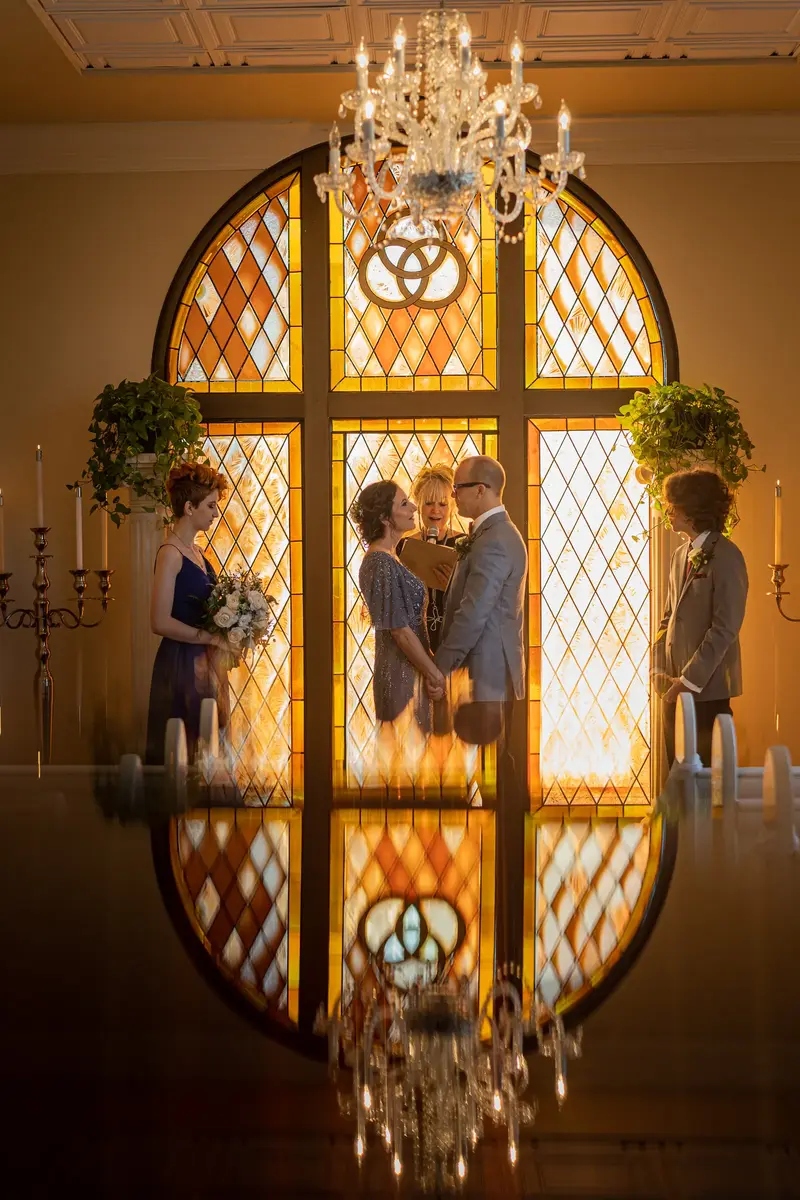 Ceremony reflected on polished floor with stained glass and chandelier above
