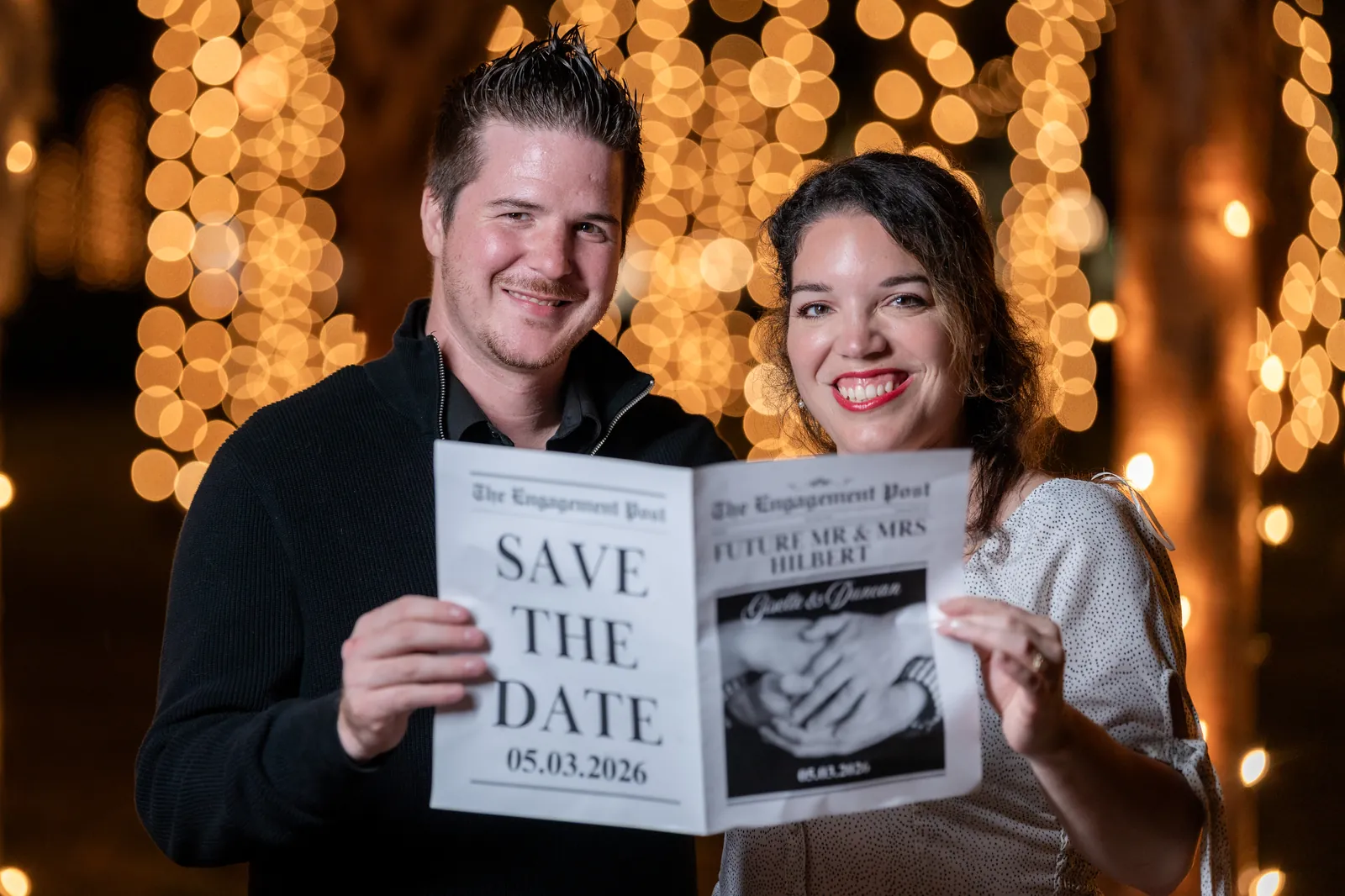 Giselle and Duncan showing save-the-date announcement with Nights of Lights bokeh