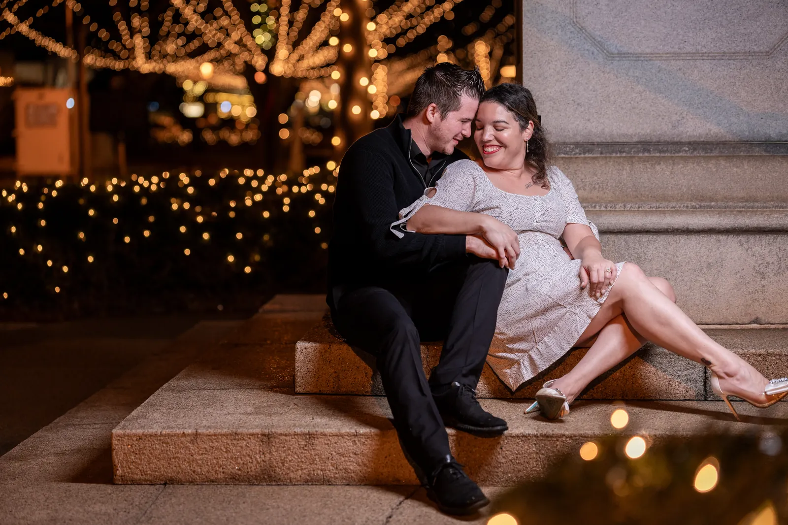 Couple cuddling on steps with Nights of Lights behind them