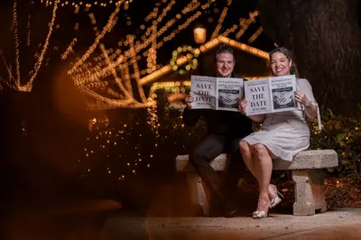 Engaged couple holding up save-the-date newspapers smiling at camera