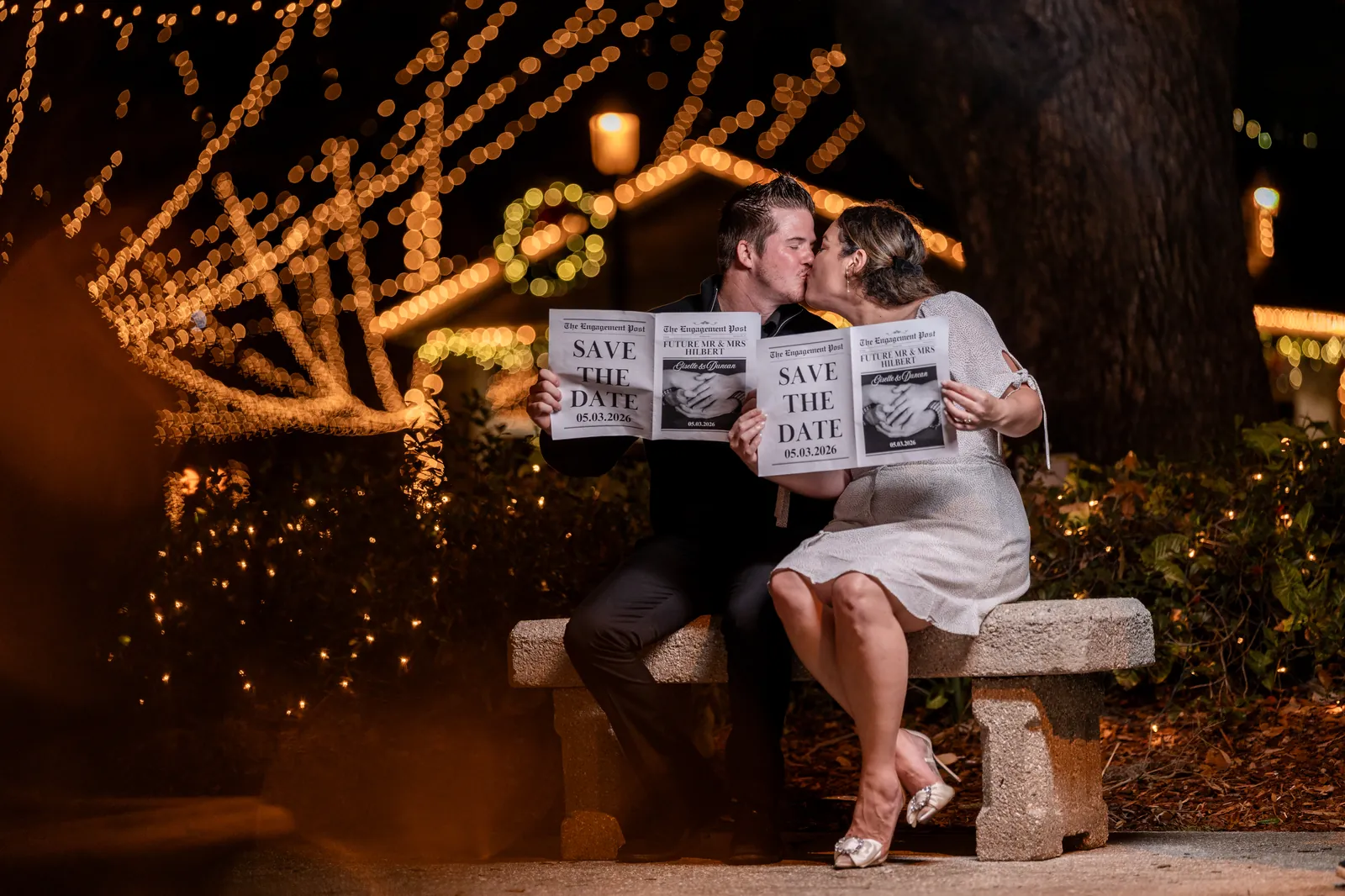 Giselle and Duncan kissing on bench with save-the-date newspapers