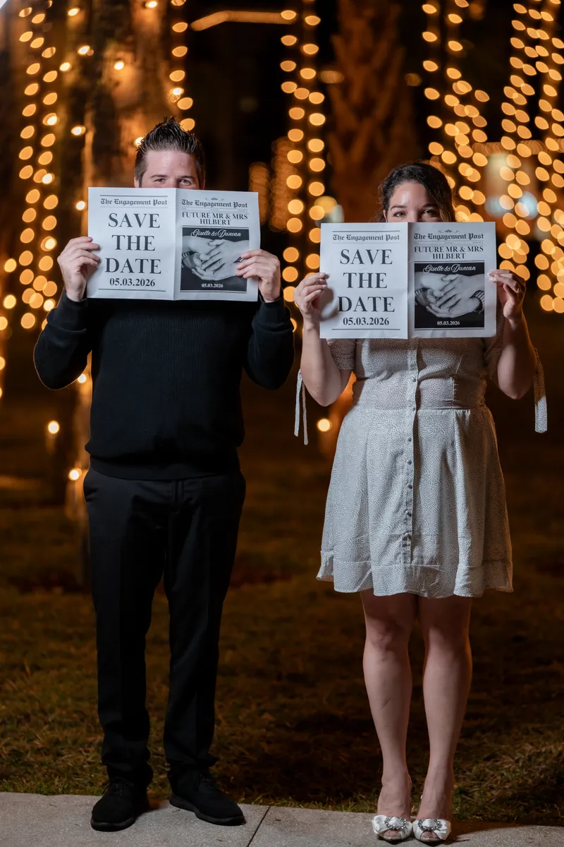 Giselle and Duncan hold up save-the-date cards in front of a glowing string-light backdrop in downtown St Augustine.