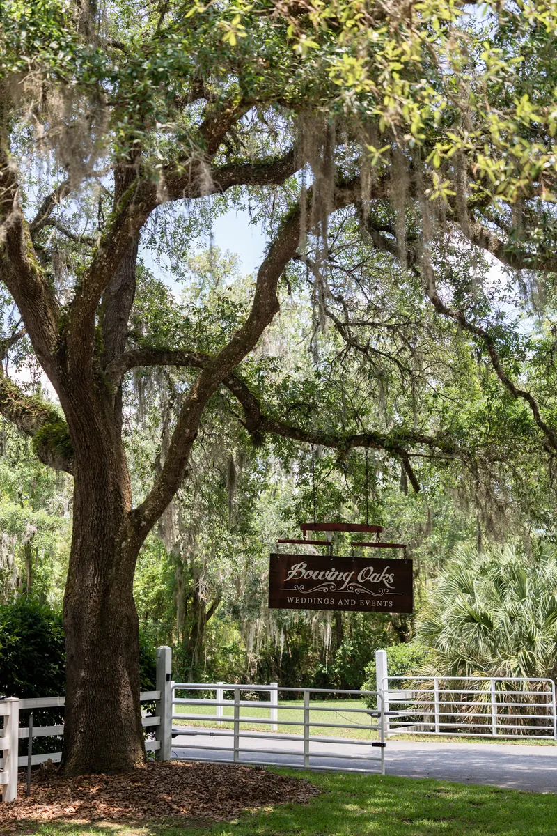 Mature oak tree with Spanish moss draping from branches, Boaring Oaks venue sign below, white fence gate entrance.