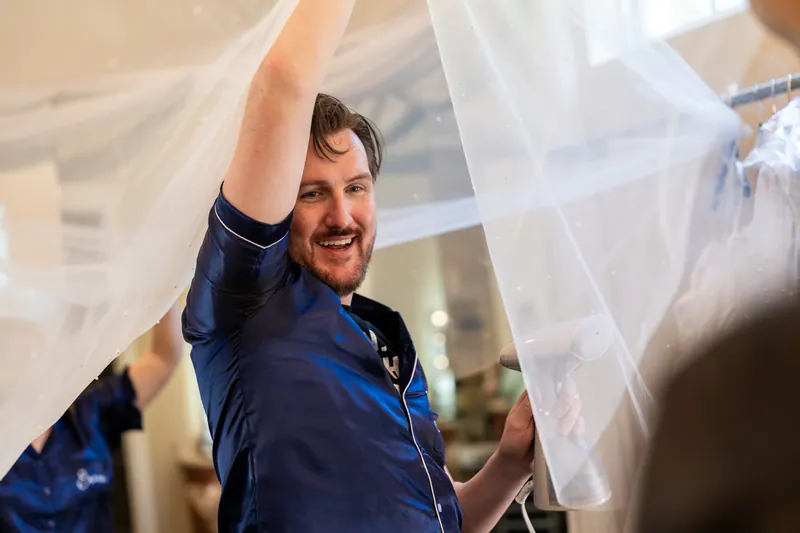 Duncan adjusting his navy suit jacket and smiling while holding his veil indoors at wedding day.