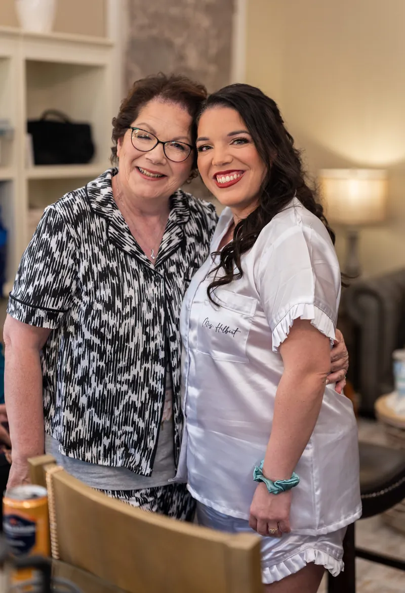 Two women embrace and smile for the camera in a bridal suite, one in a patterned shirt and one in white.