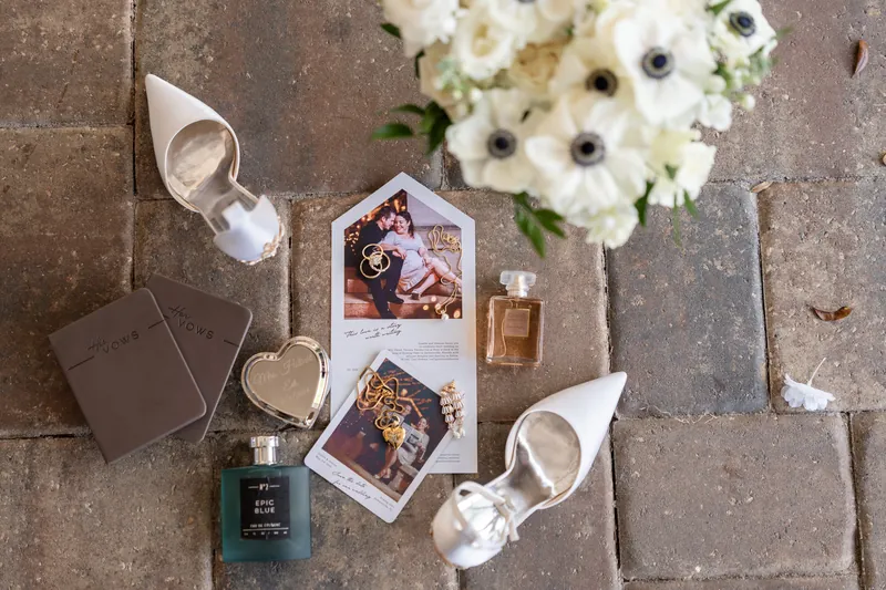 Flat lay of wedding details including white anemone flowers, perfume bottles, and framed photos on stone tile.