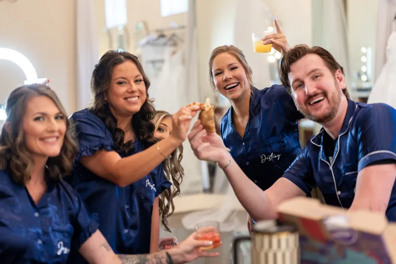Four bridesmaids in matching navy robes toast together with champagne flutes in a brightly lit room.