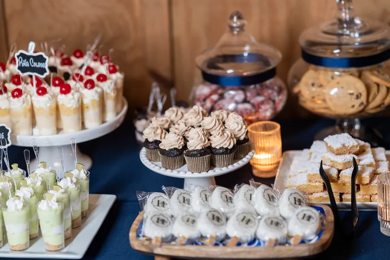 Dessert spread including green cups, berry-topped cake, chocolate cupcakes, and macarons under glass domes on blue tablecloth.
