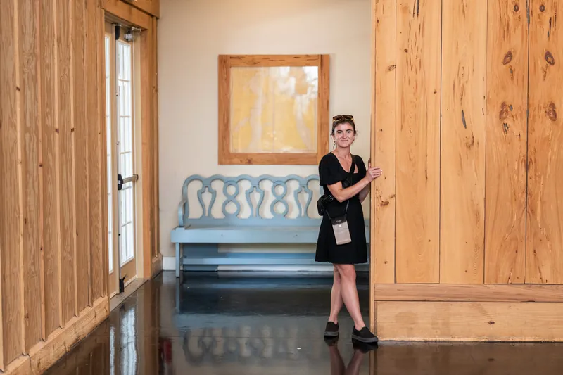 Woman in black blazer standing in wooden-walled room with gray upholstered bench and framed artwork.