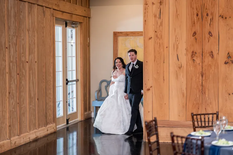 Giselle in white wedding dress and Duncan in black tuxedo walking together in wooden-paneled venue with blue tablecloth visible.