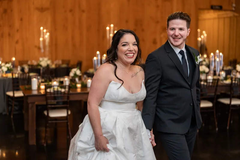 Giselle in white wedding dress and Duncan in dark suit smile together in a wood-paneled ballroom with candlelit tables.