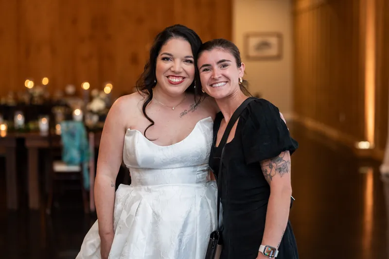 Giselle in white wedding dress stands with a woman in black, both smiling at a reception with warm lighting.