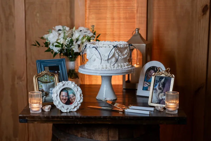 White wedding cake on pedestal with floral arrangement, framed photos, and candles on a dark table.