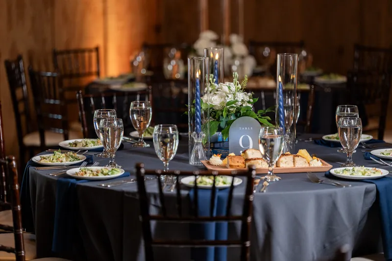 Black tablecloth with place settings, champagne glasses, white floral centerpiece, and framed photo at reception.