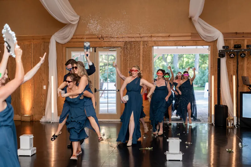 Wedding guests in navy dresses dance and celebrate on a wooden dance floor under draped fabric in wood-paneled venue.