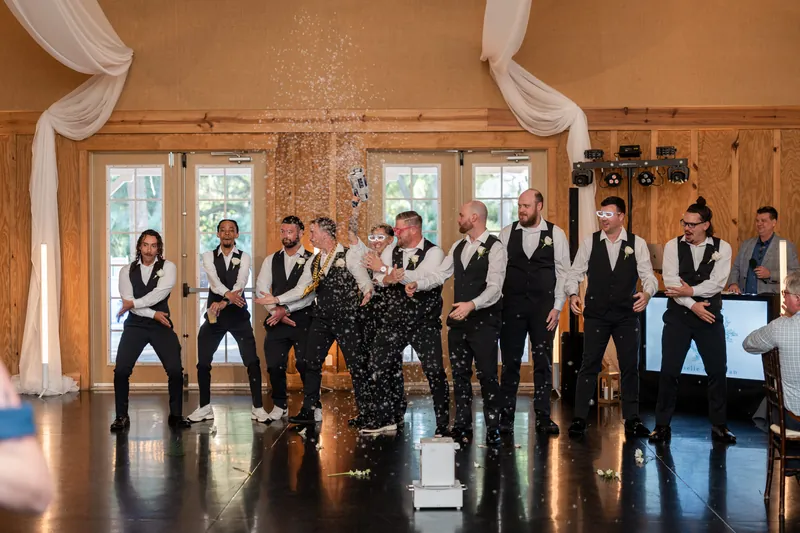 Giselle & Duncan's wedding party stands in formal attire in a wooden-beamed hall with sheer white drapes.