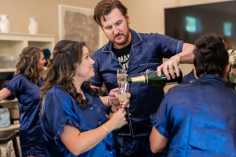 Duncan pours champagne for bridesmaids gathered around him, all wearing matching navy wedding party robes.