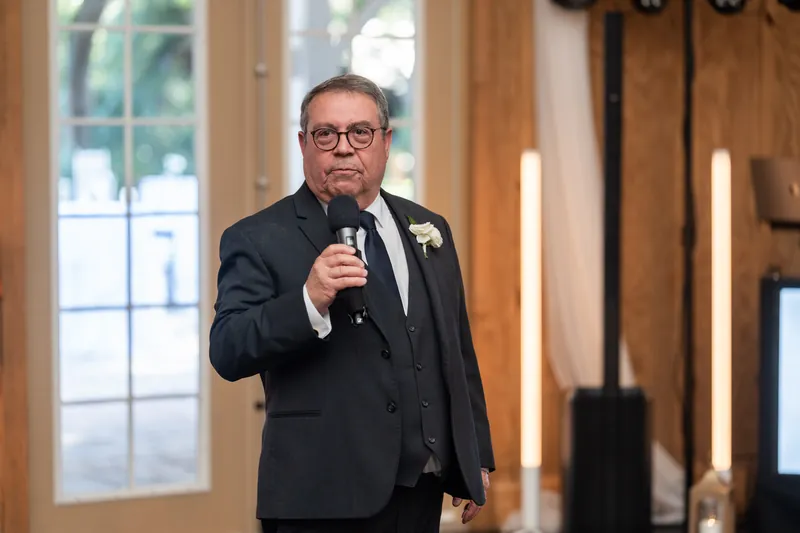 Man in dark suit and glasses holding microphone while speaking at wedding reception with white flowers pinned to lapel.