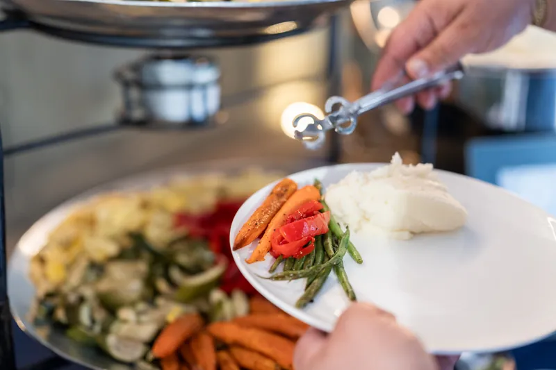 Server plating seared scallop with vegetables using tongs over white plate.