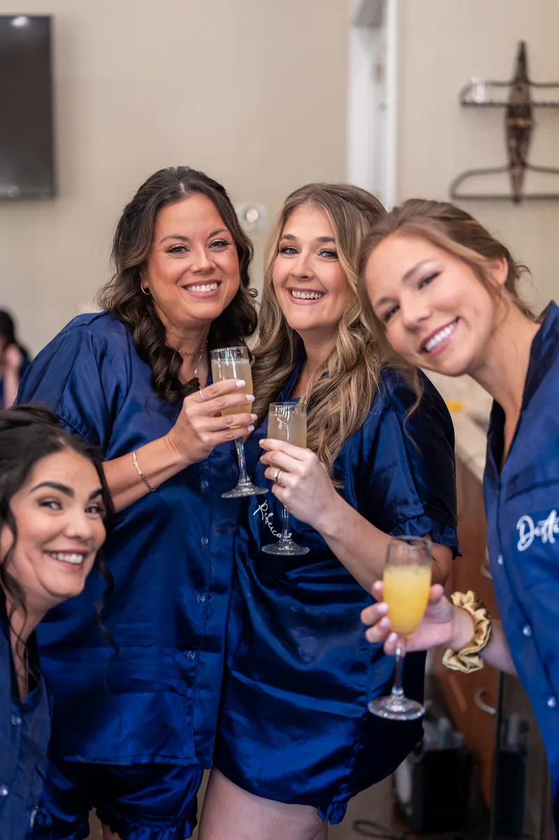 Giselle and three bridesmaids clink champagne glasses together, laughing in their navy silk robes.