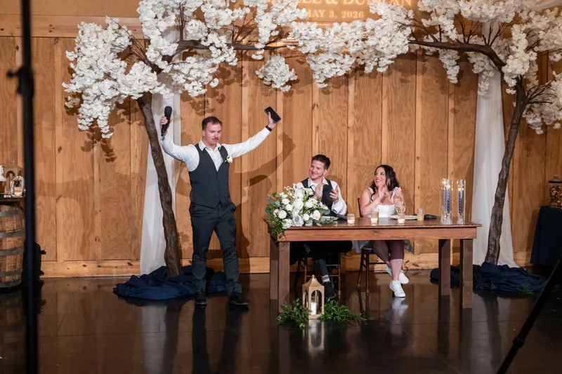 Duncan at altar with arms raised under white floral arch while Giselle and officiant sit at wooden table.