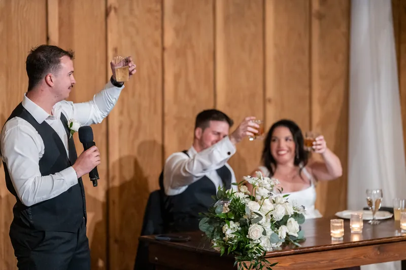 Best man raises his glass while Duncan and Giselle give thumbs up during reception toast at wooden venue.
