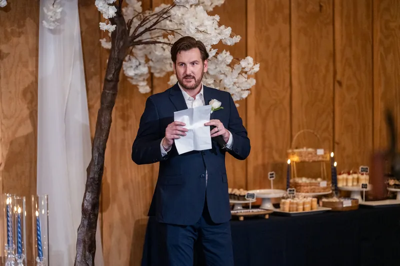 Duncan holds note cards while speaking at wedding reception with white flowering branch decoration behind him.