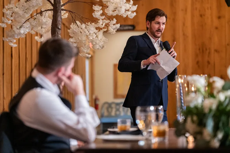 Duncan speaks at reception while guest listens, with white flowering branch and dinner table in foreground.