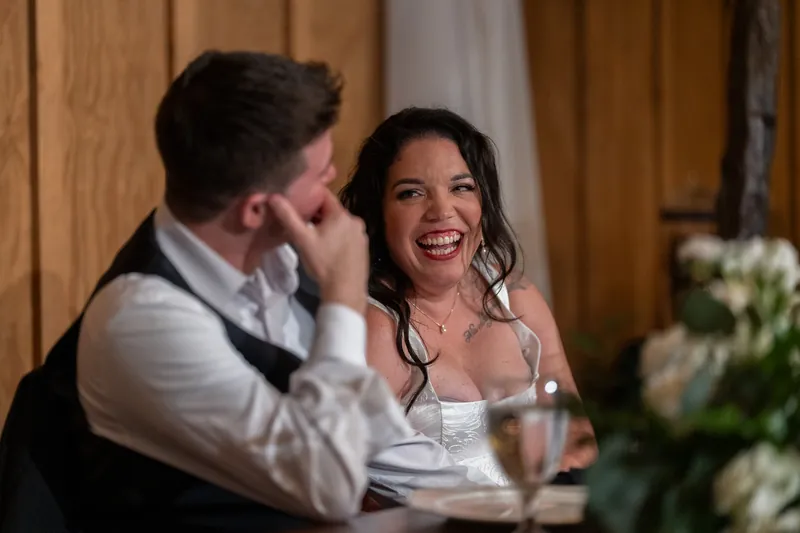 Duncan and Giselle smile at each other during reception dinner, seated at table in wooden hall.