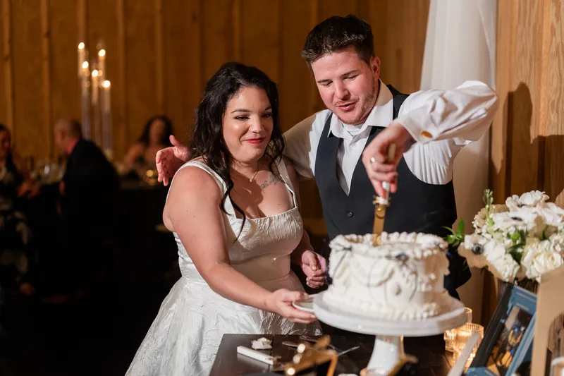 Giselle and Duncan cut their white wedding cake together at reception, laughing with guests in background.