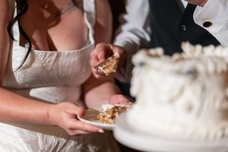Giselle and Duncan hold plates with cake slices, standing beside their tiered white wedding cake.