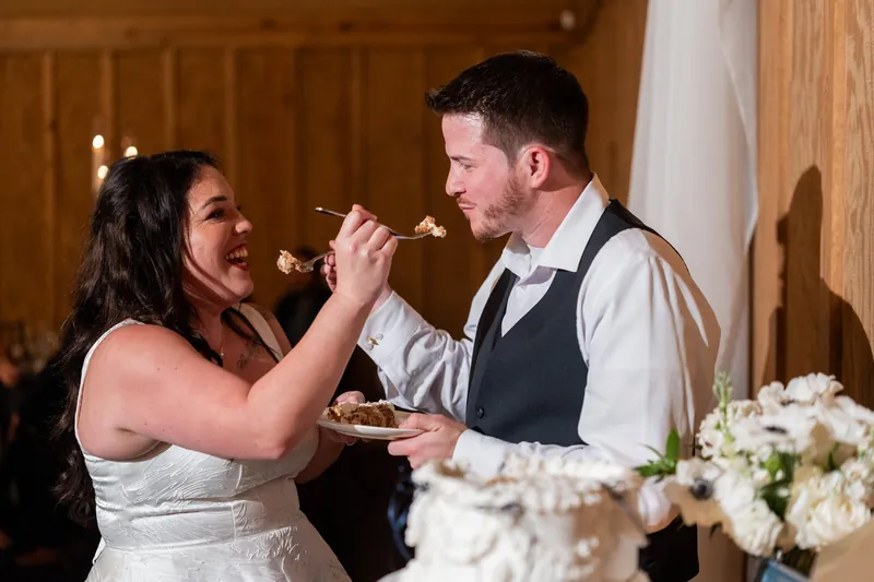 Giselle feeds Duncan cake during their reception in a wood-paneled hall with white floral centerpieces nearby.