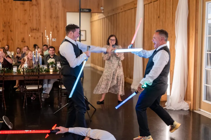 Two groomsmen engage in a light saber battle while a woman watches, surrounded by seated guests in the wooden venue.