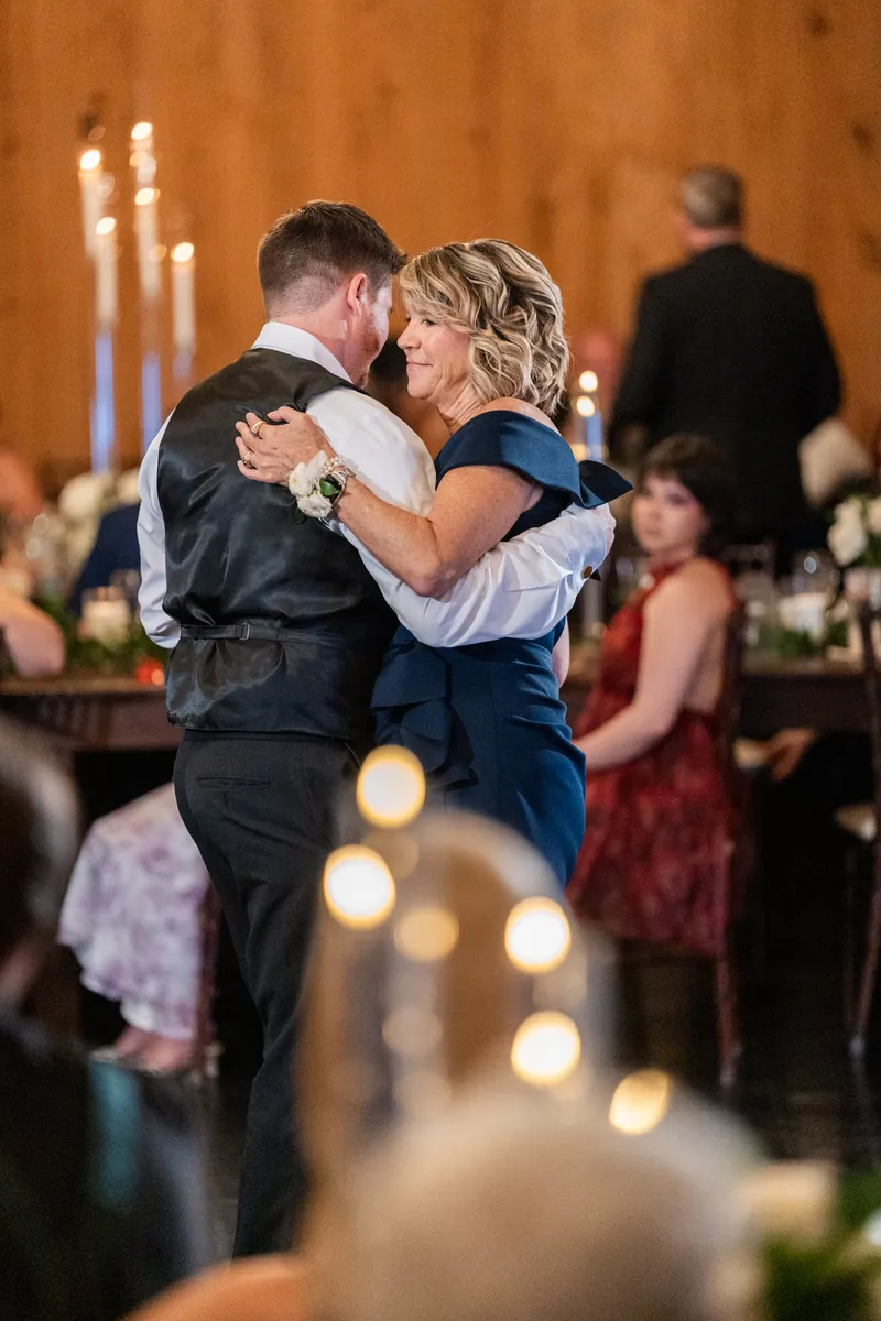 Duncan and Giselle embrace on the dance floor during their reception in a wood-paneled venue with candles and white flowers.