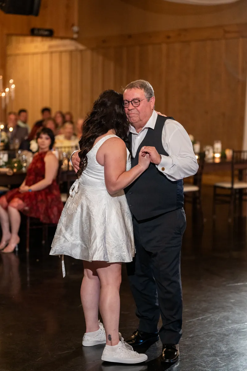 Giselle dances with her father in a white dress while guests watch in the wooden reception hall.