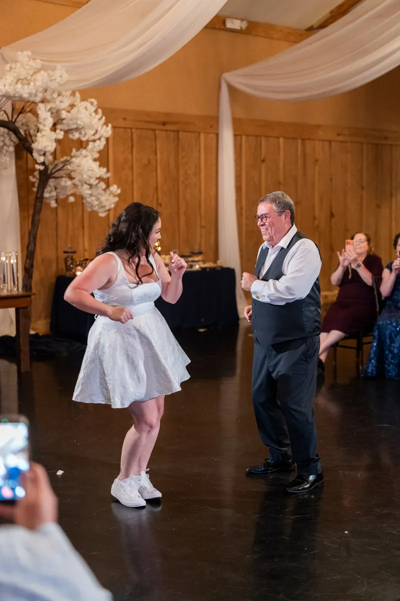 Giselle dances with her father in a white dress on the wooden dance floor as guests applaud.