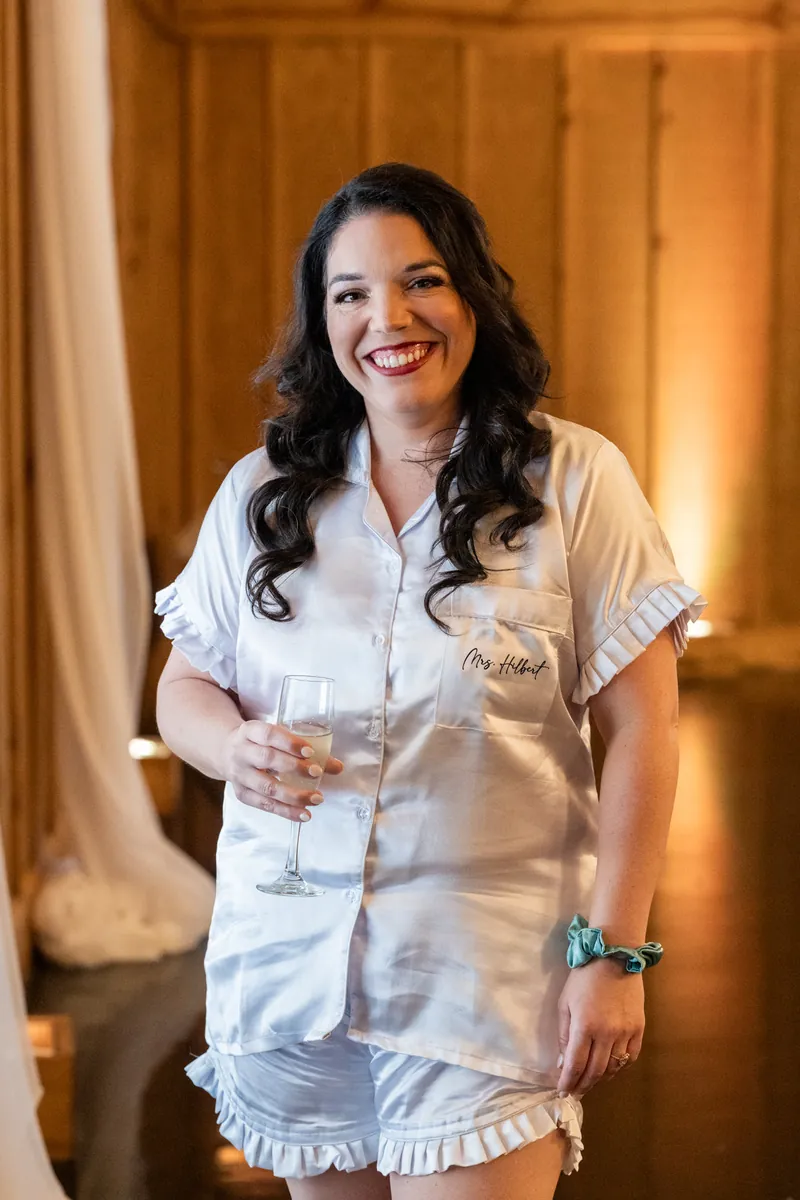 Giselle smiles while holding a drink in a white button-up shirt inside a warm-lit wooden room.